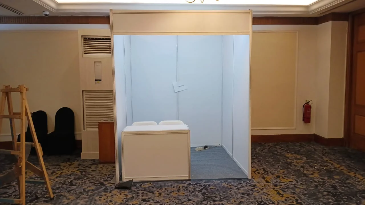 Booth pameran minimalis berpanel putih dengan counter depan untuk acara konferensi di ballroom
