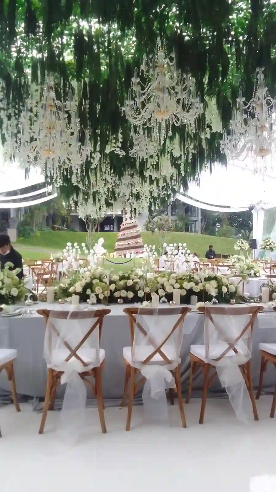 Meja pengantin panjang dengan kursi crossback bersash tulle putih dan rangkaian bunga putih di tenda transparan oleh Eventara Indonesia