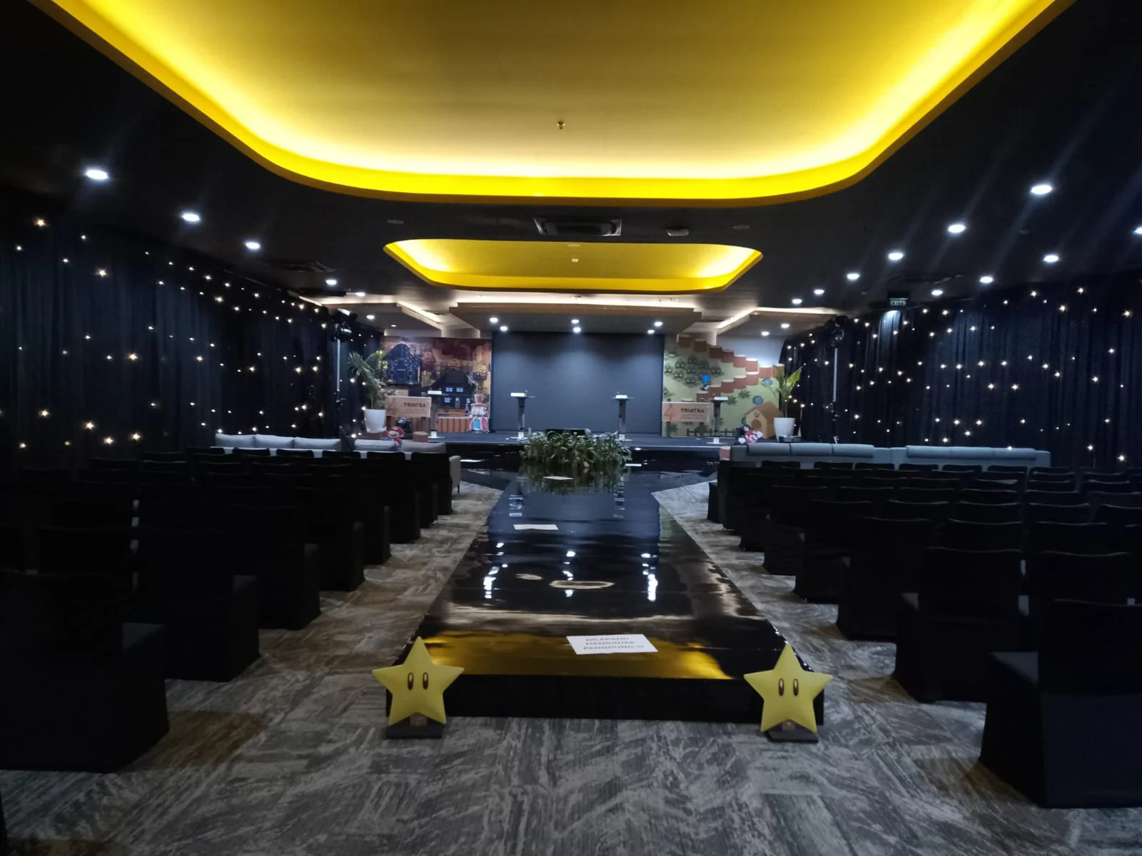 Ballroom Triatra Fest dengan susunan kursi menghadap panggung, runway hitam dan dekorasi retro gaming
